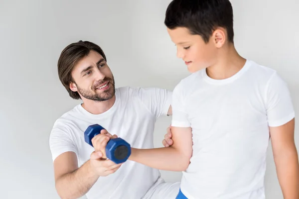 Padre ayudando a su hijo pequeño con mancuerna a ejercitarse sobre fondo gris - foto de stock