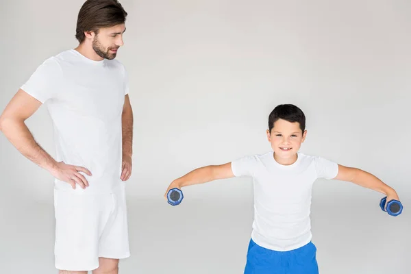 Man standing akimbo and looking at son with dumbbells exercising on grey backdrop — Stock Photo