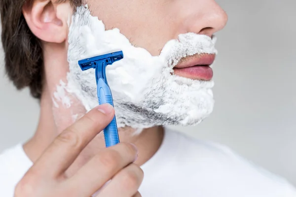 Cropped shot of man with razor in hand and shaving foam on face on grey backdrop — Stock Photo