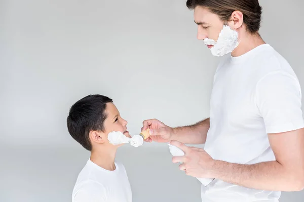 Vista lateral del padre con cepillo poniendo espuma de afeitar en la cara hijos sobre fondo gris - foto de stock