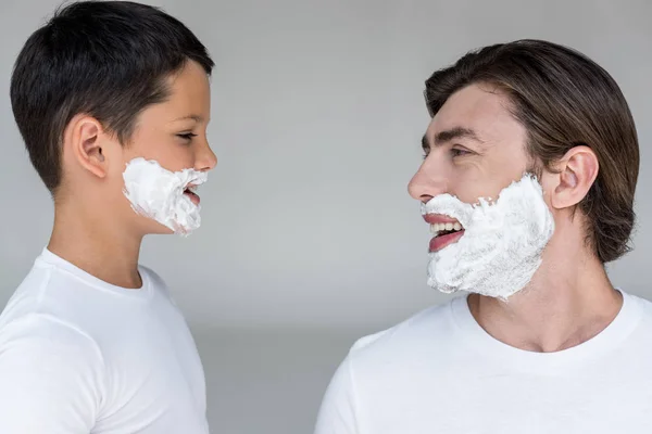 Visão lateral de pai e filho feliz com espuma de barbear em rostos no fundo cinza — Fotografia de Stock