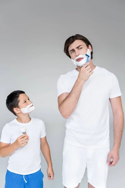 Portrait of father and son with shaving foam on faces with brush and razor in hands on grey background — Stock Photo