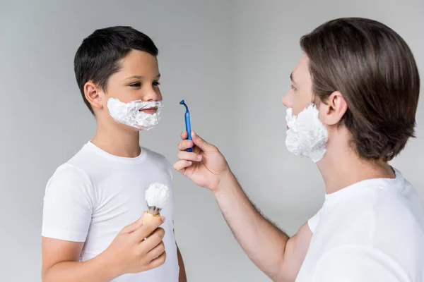 Father and son with shaving foam on faces with brush and razor in hands isolated on grey — Stock Photo