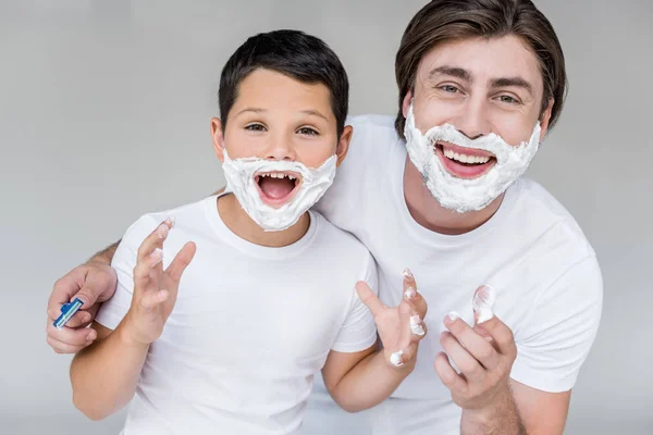 Портрет эмоционального отца и сына с пеной для бритья на лицах, изолированных на сером — стоковое фото