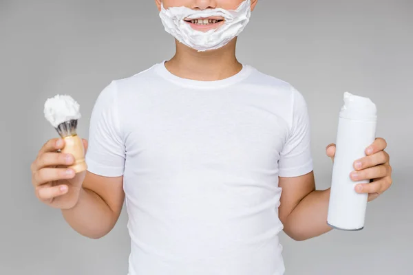 Partial view of smiling preteen boy with brush, cream and shaving foam on face isolated on grey — Stock Photo