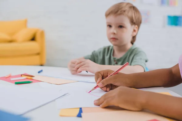 Partial view of multicultural preschoolers at table with papers and pencils in classroom — Stock Photo