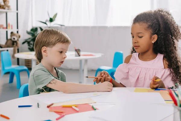 Portrait of multicultural preschoolers at table with papers and pencils in classroom — Stock Photo