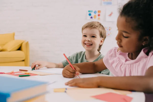 Focus selettivo di bambini in età prescolare multiculturale a tavola con carte e matite in classe — Foto stock