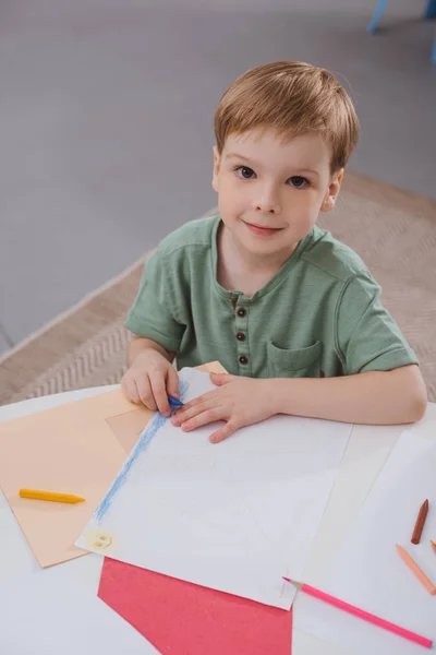 High angle view of preschooler boy sitting at table with paper and colorful pencils for drawing in classroom — Stock Photo