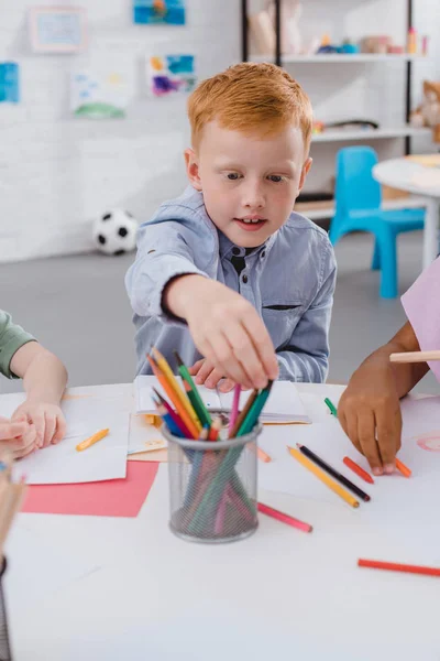 Selective focus of red hair boy taking pencil at table with multicultural classmates in classroom — Stock Photo