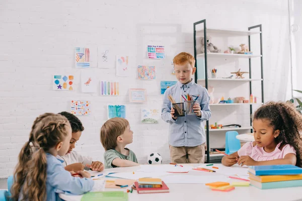 Interracial kids sitting at table with papers and paints for drawing in classroom — Stock Photo