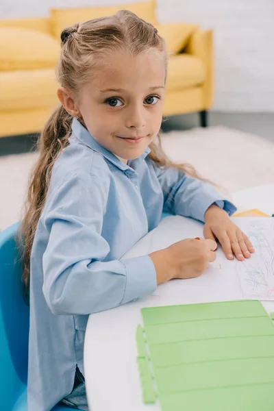 Vista lateral del niño sonriente con lápiz sentado en la mesa con papeles para dibujar - foto de stock