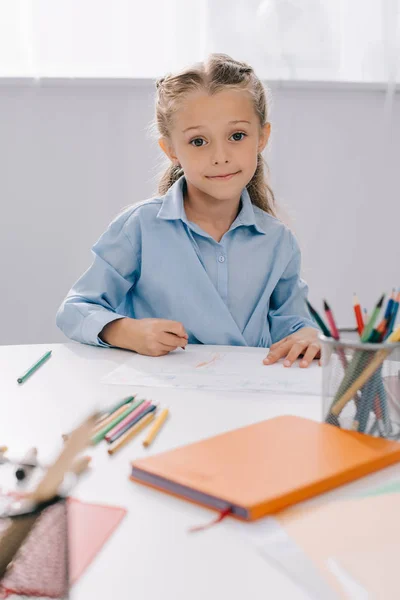 Retrato de niño sonriente sentado en la mesa con lápices de colores y papeles para dibujar - foto de stock