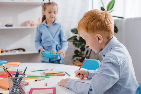 Enfoque selectivo de pelo rojo niño dibujo imagen mientras compañero de clase de pie cerca de la mesa - foto de stock