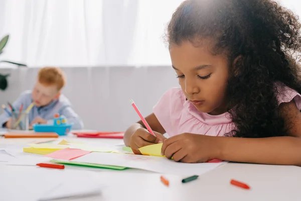 Focus selettivo di bambini in età prescolare multiculturale che disegnano immagini con matite in classe — Foto stock