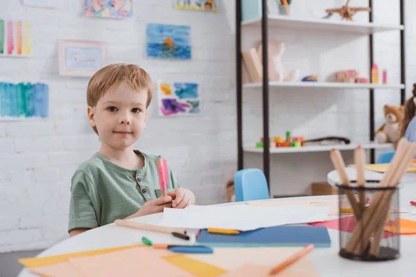 Portrait of preschooler boy sitting at table with paper and colorful pencils for drawing in classroom — Stock Photo