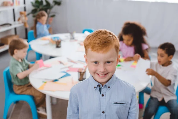 Foco seletivo de menino cabelo vermelho feliz olhando para a câmera com colegas multirraciais atrás em sala de aula — Fotografia de Stock