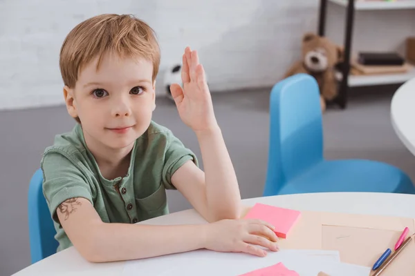Портрет милого дошкольника с поднятой рукой, сидящего за столом в классе — стоковое фото