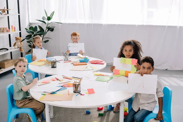 Niños multiétnicos mostrando fotos mientras están sentados en la mesa en el aula - foto de stock