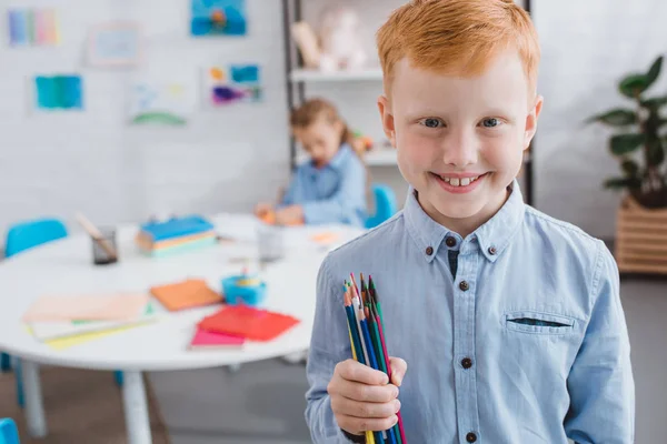 Enfoque selectivo de niño pelo rojo feliz con lápices y compañero de clase en la mesa en el aula - foto de stock