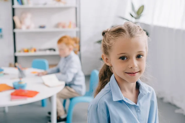 Selektiver Fokus eines lächelnden Vorschülers, der in die Kamera blickt, mit einem Mitschüler am Tisch im Klassenzimmer — Stockfoto