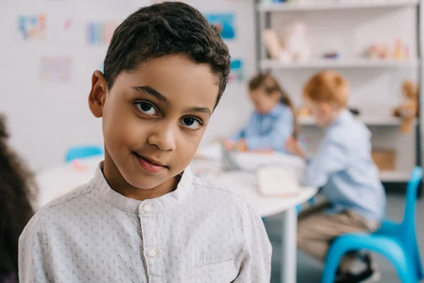 Selektiver Fokus des süßen afrikanisch-amerikanischen Jungen, der mit Klassenkameraden im Klassenzimmer in die Kamera schaut — Stockfoto