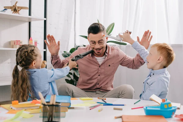 Teacher in eyeglasses with hands up and kids with toy guns playing in classroom — Stock Photo