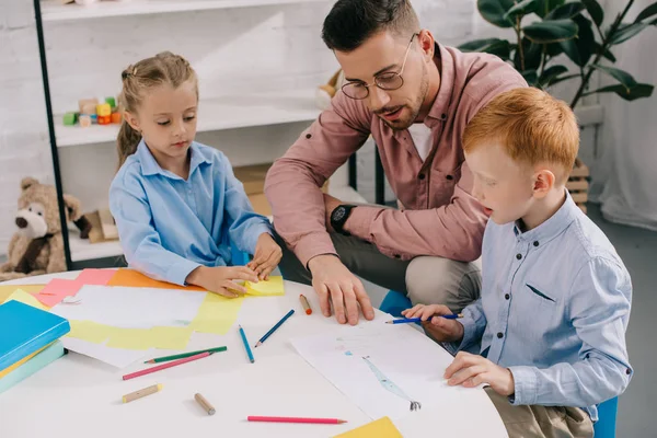 Insegnante in occhiali aiutare i bambini in età prescolare con il disegno a tavola in classe — Foto stock