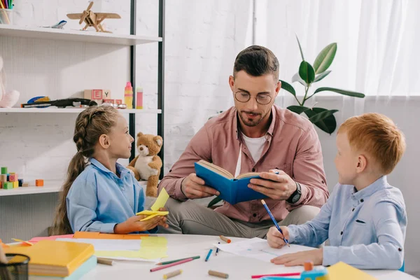 Profesor en anteojos lectura libro a los niños en la mesa en el aula — Stock Photo