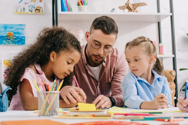 Insegnante di occhiali che aiuta i bambini in età prescolare multiculturale con il disegno a tavola in classe — Foto stock