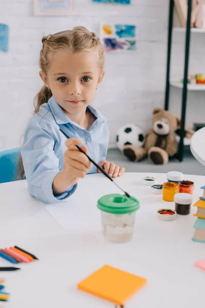 Portrait of cute child sitting at table with paints and paint brush — Stock Photo