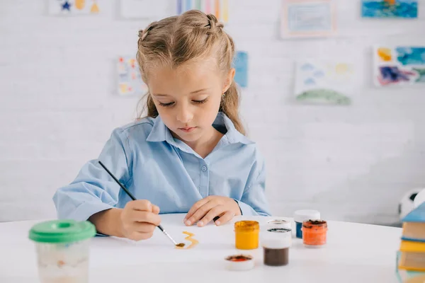 Портрет очаровательной сфокусированной детской рисунки с красками и кистью за столом — стоковое фото