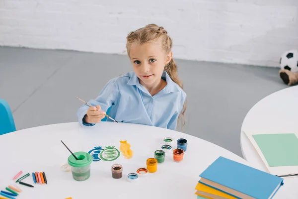 Портрет милого ребенка, сидящего за столом с красками и кистями — стоковое фото