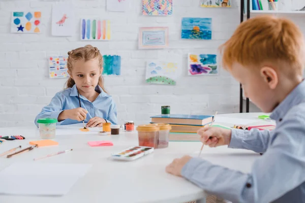 Enfoque selectivo de los niños en edad preescolar dibujos con pinturas y pinceles en la mesa en el aula - foto de stock