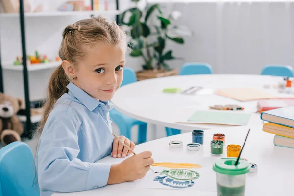 Vista lateral del niño lindo sentado en la mesa con pinturas y pinceles - foto de stock