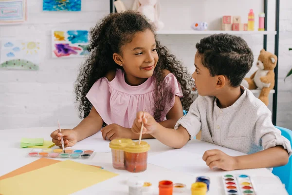 Niños afroamericanos mientras se miran los unos a los otros haciendo dibujos con pinturas y pinceles en el aula - foto de stock