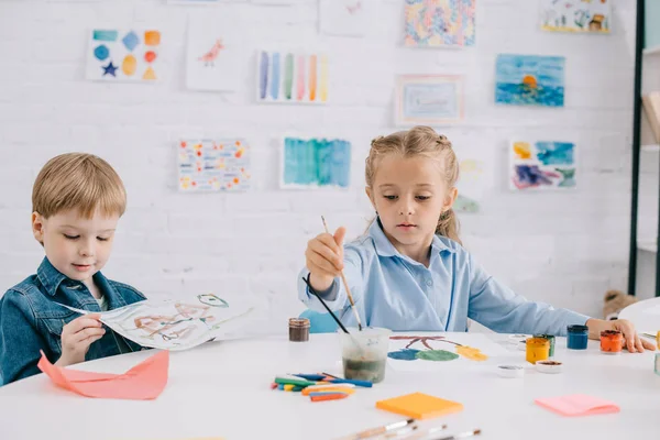 Niños preescolares enfocados dibujando cuadros con pinturas y pinceles en la mesa en el aula - foto de stock