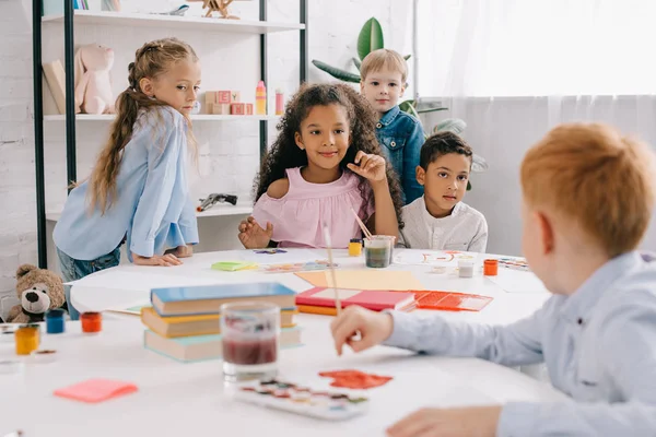 Messa a fuoco selettiva di bambini multietnici con pennelli disegno immagini in classe — Foto stock