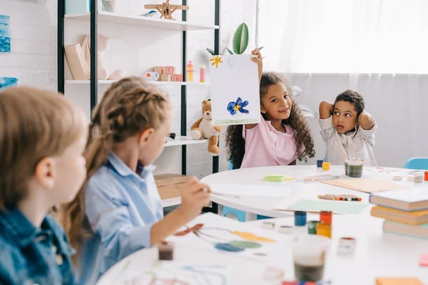 Focus selettivo del bambino afroamericano che mostra foto ai compagni di classe multiculturali in classe — Foto stock