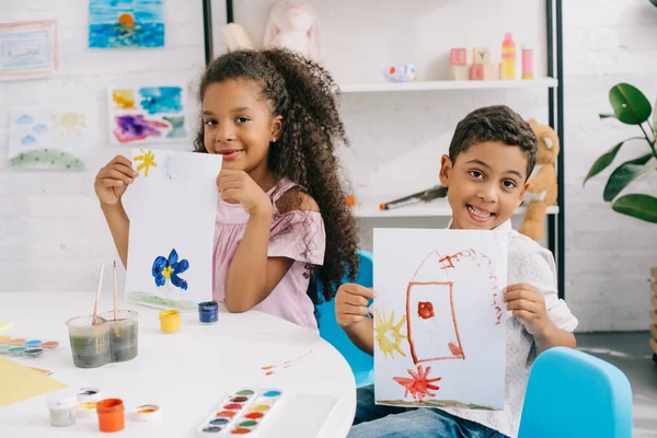 Retrato de niños afroamericanos felices mostrando fotos en la mesa en el aula - foto de stock