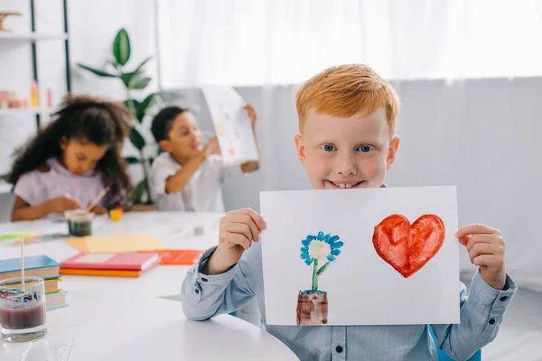 Selective focus of smiling red hair boy showing picture in hands with african american classmates behind in classroom — Stock Photo