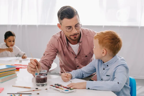 Вчителька допомагає маленькому хлопчику малювати картину за столом у класі — стокове фото