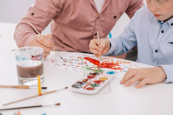 Частковий вид вчителя, який допомагає маленькому хлопчику малювати картину за столом у класі — стокове фото