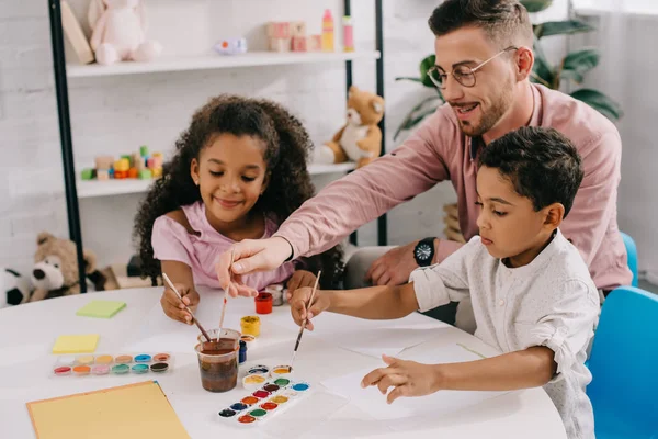 Sorridente professor caucasiano em óculos e crianças afro-americanas desenhando quadros com tintas juntas em sala de aula — Fotografia de Stock