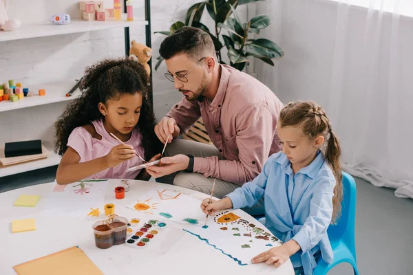 Lehrer mit Brille und multiethnische Kinder malen Bilder mit Farben am Tisch im Klassenzimmer — Stockfoto