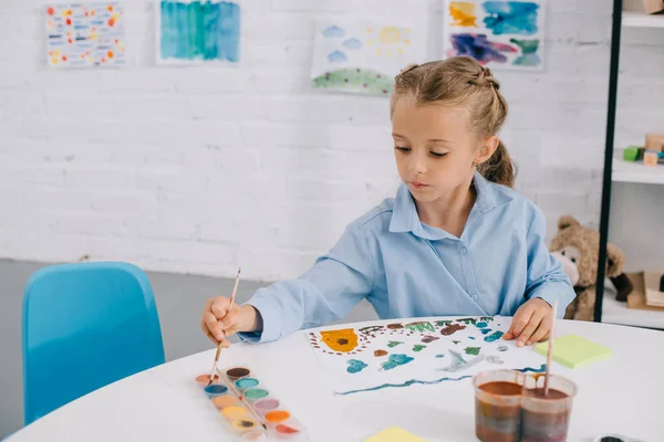 Retrato de adorável criança focada desenho colorido quadro com tintas e escova à mesa — Fotografia de Stock