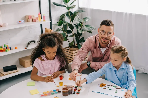Lehrer mit Brille und multiethnische Kinder malen Bilder mit Farben am Tisch im Klassenzimmer — Stockfoto
