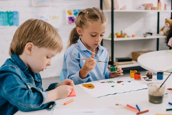 Des enfants d'âge préscolaire concentrés dessinant des images avec des peintures et des pinceaux à table en classe — Photo de stock