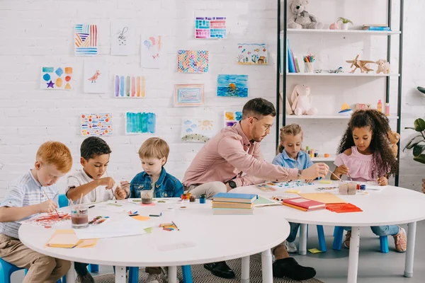 Enseignant et interracial enfants dessin images ensemble à tables dans salle de classe — Photo de stock