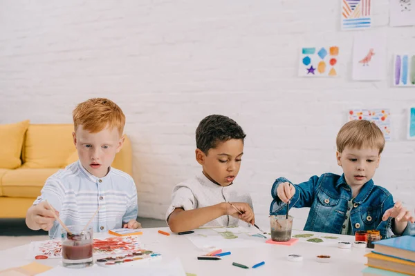 Retrato de niños multiétnicos con pinceles pintando dibujos en el aula - foto de stock